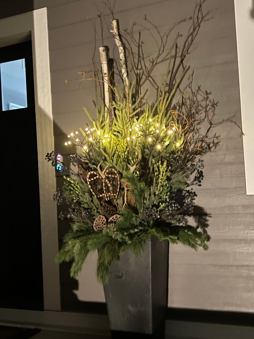 Elegant holiday arrangement in a tall planter with greenery, twigs, and soft glowing lights.