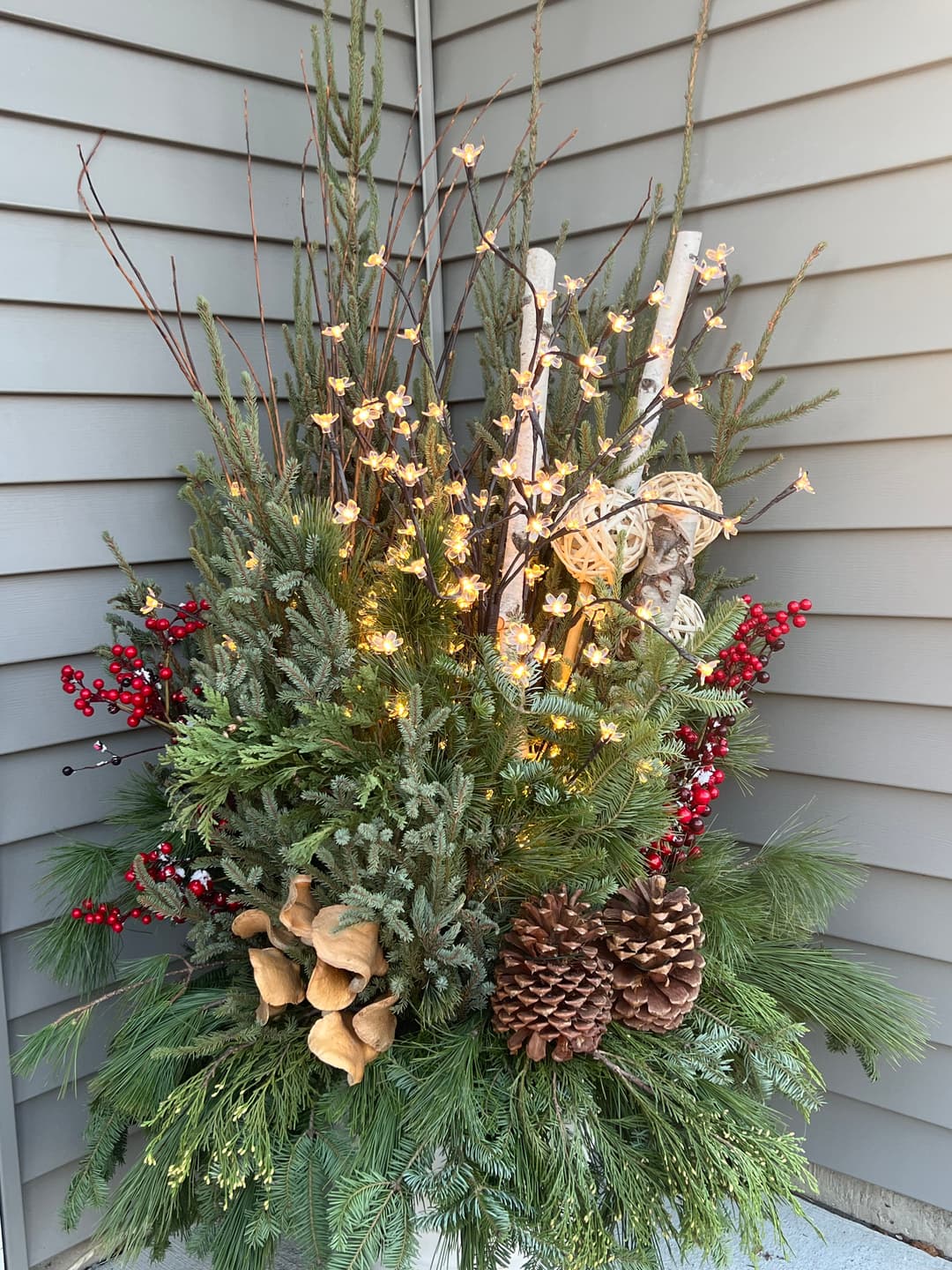 Festive outdoor winter arrangement with pinecones, berries, and twinkling lights.