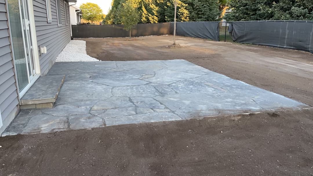 Stone patio installation in a spacious backyard with a gravel border and landscape.