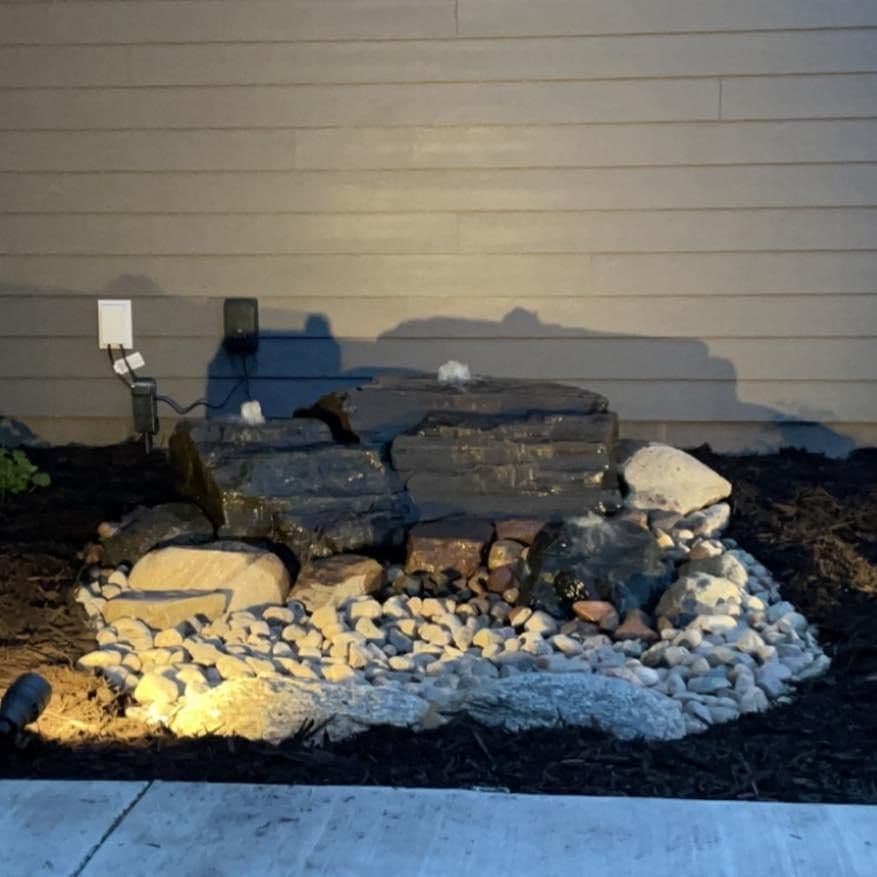 Landscape feature with rocks, pebbles, and illuminated water fountain against a house wall.