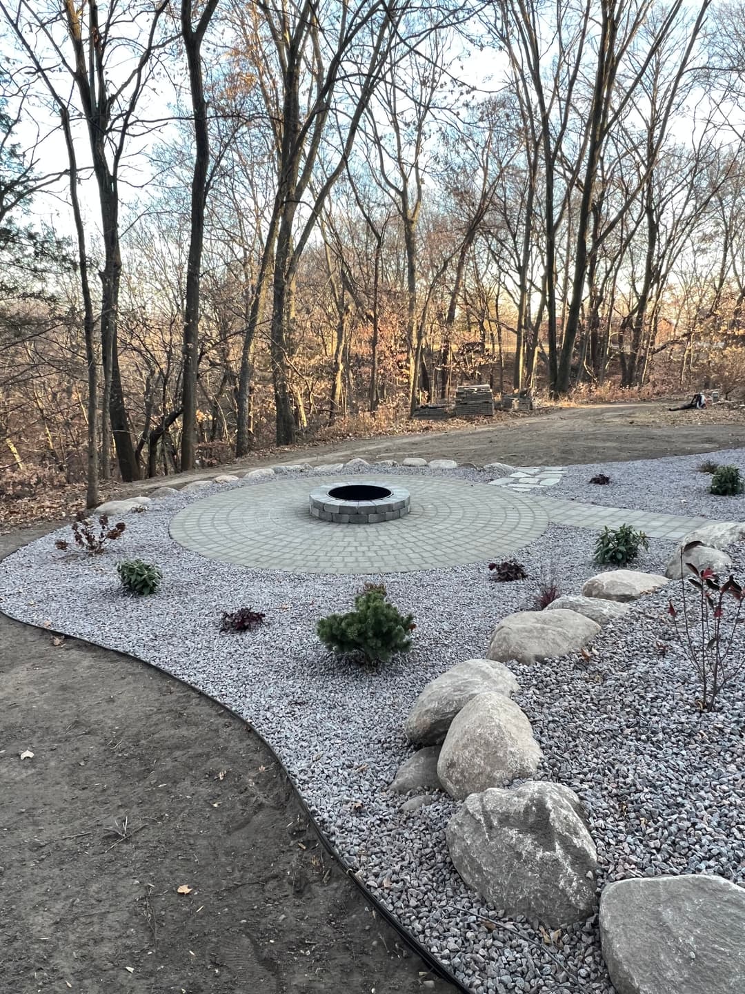 Fire pit surrounded by gravel and plants in a forested area during sunset.