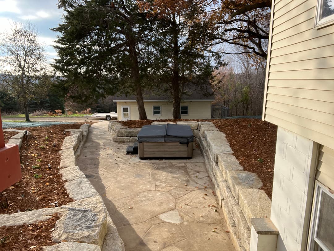 Hot tub nestled in a landscaped patio area with stone walls and trees in the background.