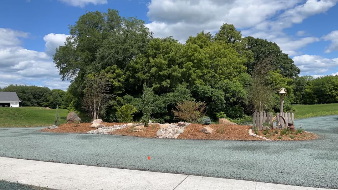 Landscape design featuring plants, mulch, rocks, and a decorative fence under a cloudy sky.