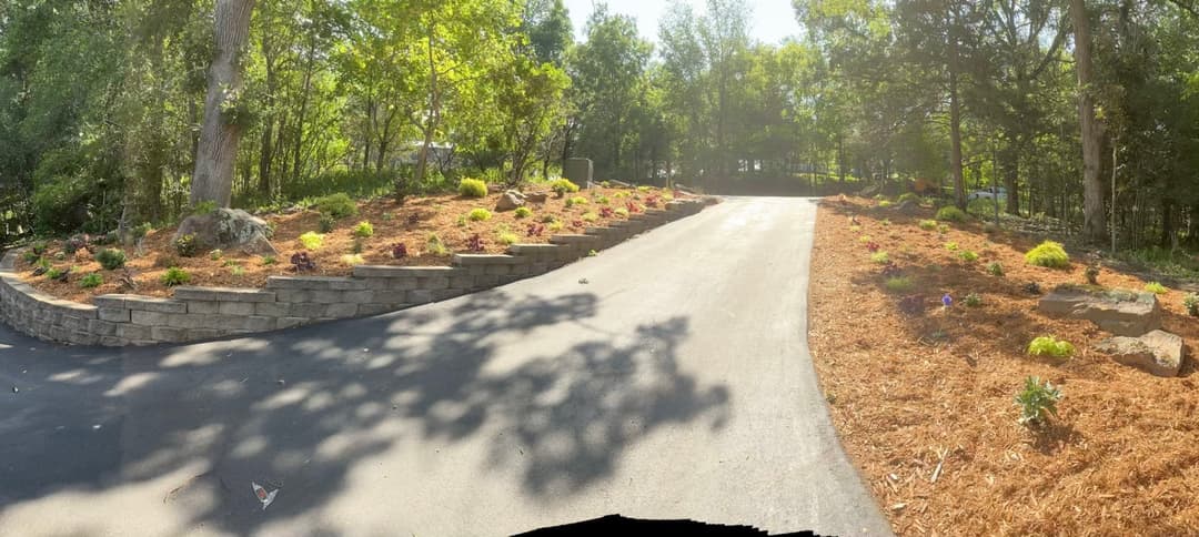 Landscaped driveway with stone wall, vibrant flowers, and mulched garden beds surrounded by trees.