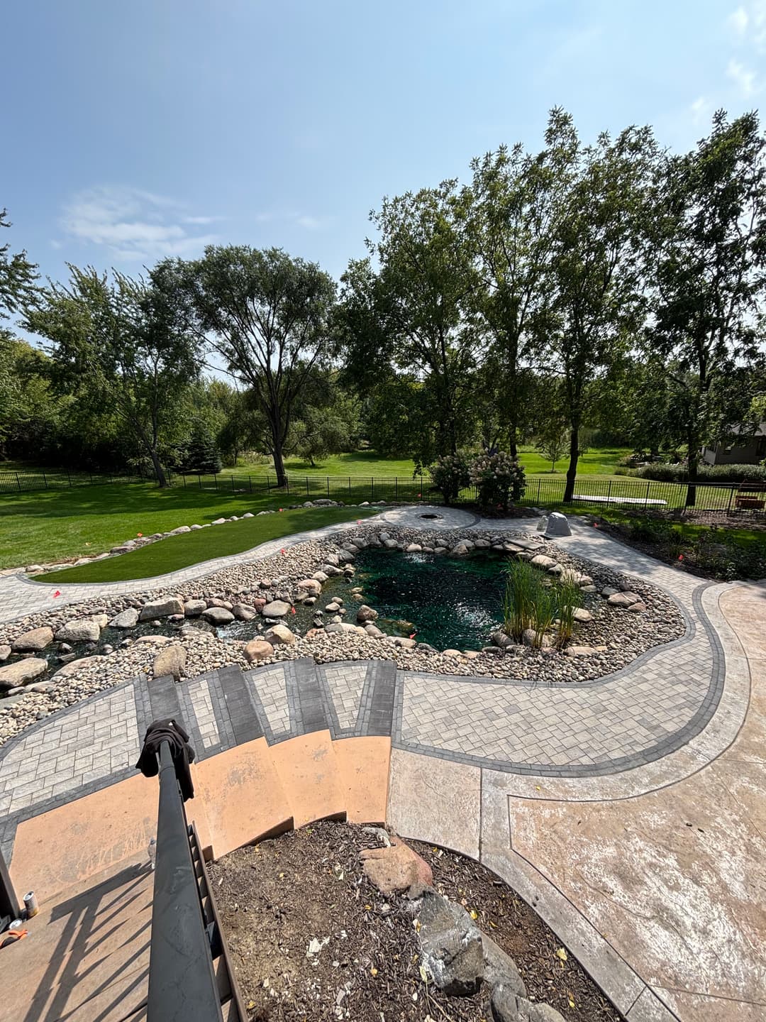 Scenic backyard with a pond, landscaped stones, and lush greenery under clear blue sky.