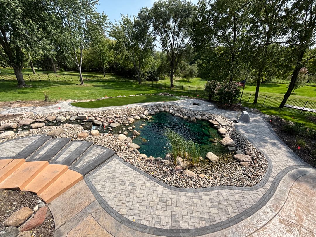 Serene backyard landscape featuring a pond, stones, and paved walkway surrounded by greenery.