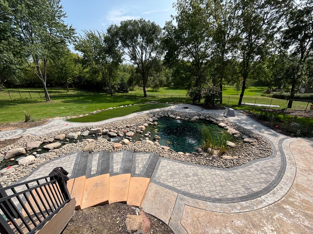 Scenic backyard pond with rocks, foliage, and patio area in a lush green landscape.