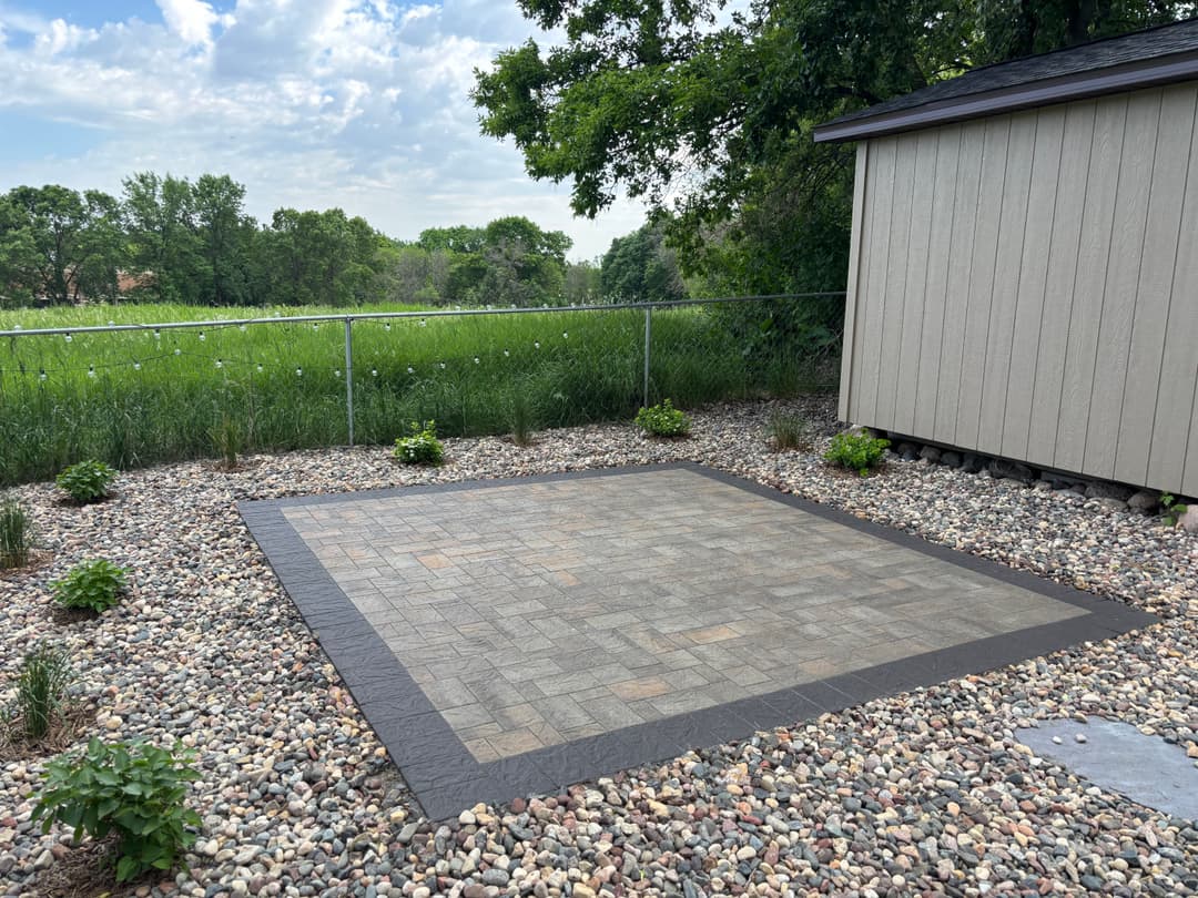Patio area with pavers surrounded by landscaping and rocks near a shed and grassy field.
