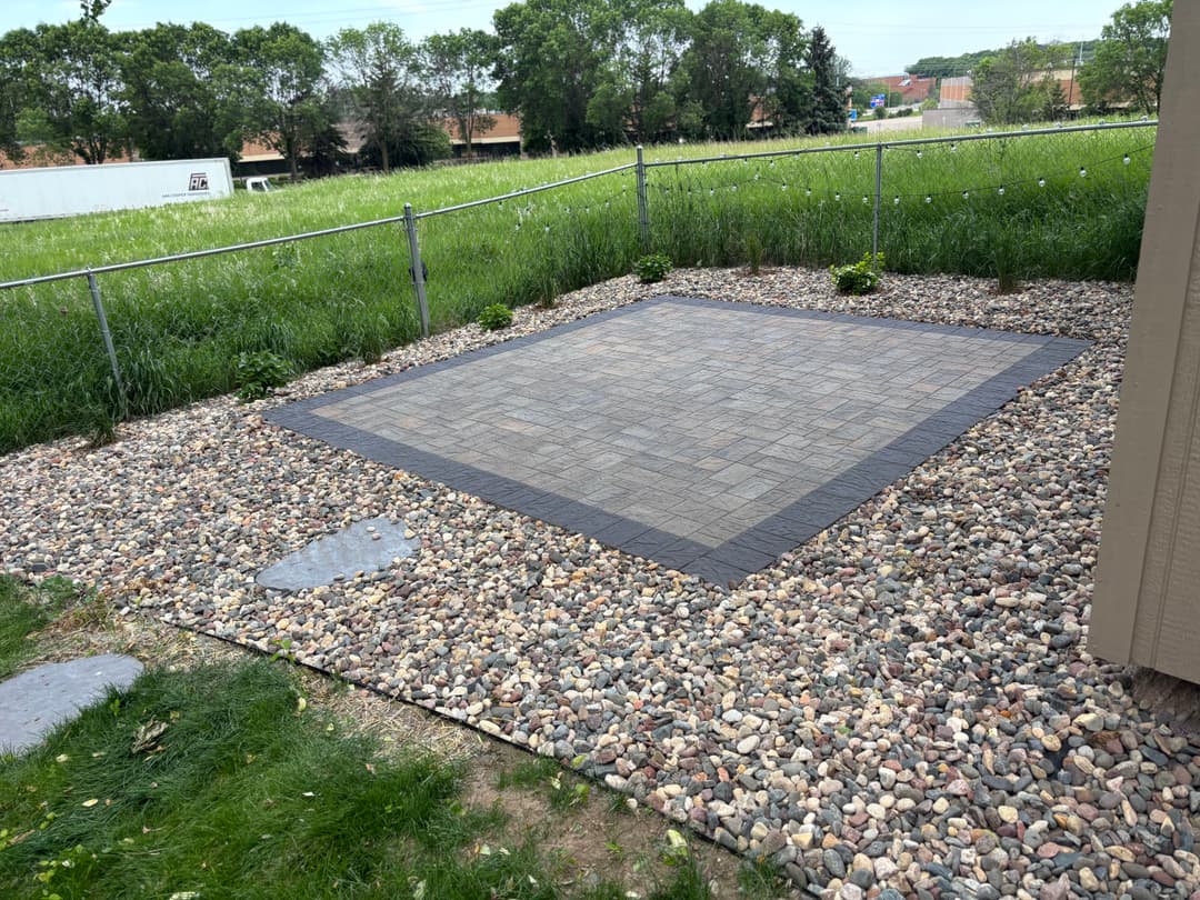Paver stone patio surrounded by decorative gravel and greenery in a fenced yard.