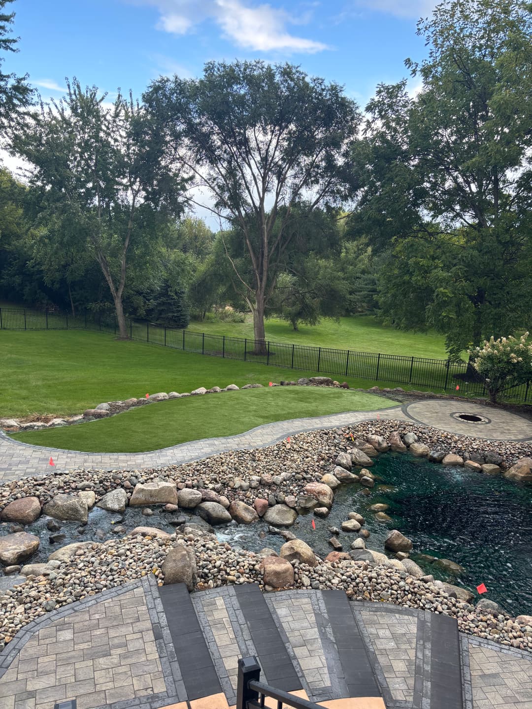 Lush green backyard with landscaped pond, stone pathways, and vibrant trees under a blue sky.