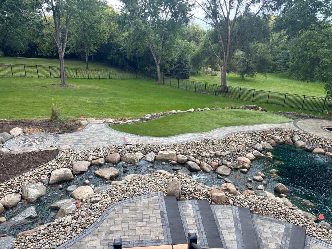 Lush backyard landscape with a stone pathway, pond, and manicured grass surrounded by trees.