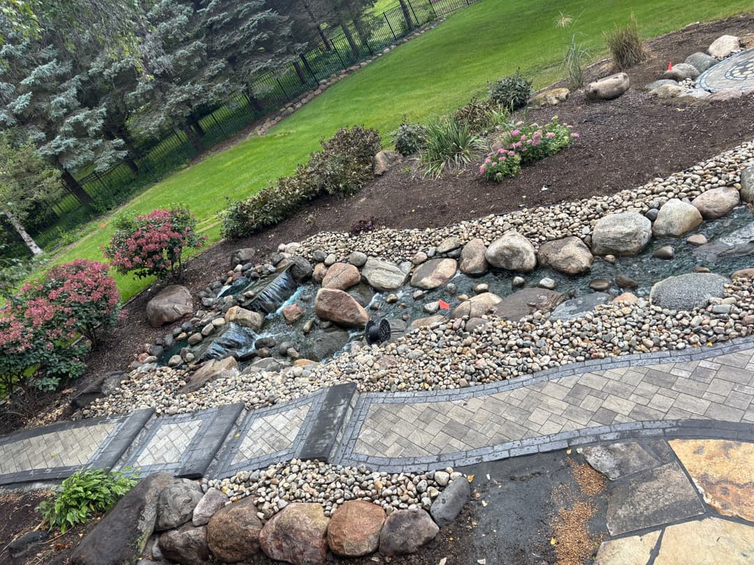 landscaped garden featuring a winding stone path, water stream, and lush greenery.