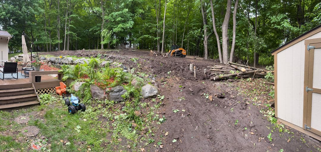 Cleared backyard with muddy terrain, trees, and landscaping equipment in forested area.